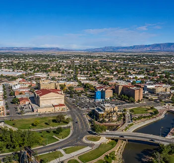 Aerial view of Pueblo, Colorado, with downtown buildings, a riverfront bridge, and mountain views in the background.