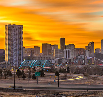 Denver skyline at sunset with glowing orange sky, city buildings, and a blue bridge over a highway.