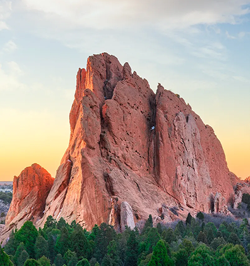 Large red sandstone rock formation at sunset with green trees in the foreground and a soft cloudy sky above.